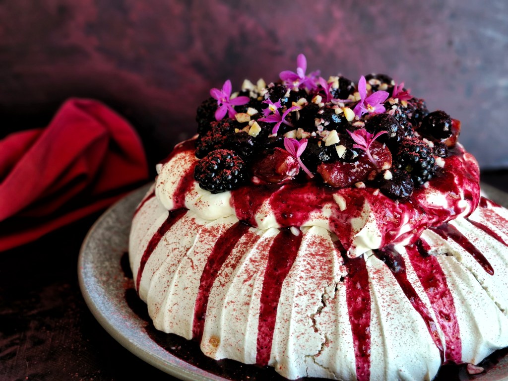 Native plum pavlova topped with dark berries, plum sauce and pink edible flowers.